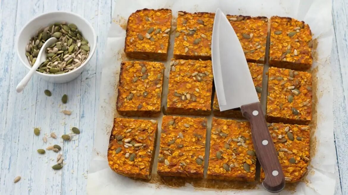 Tray of pumpkin-seed bars cut into squares on parchment, with a knife resting on top and a bowl of mixed seeds to the side.