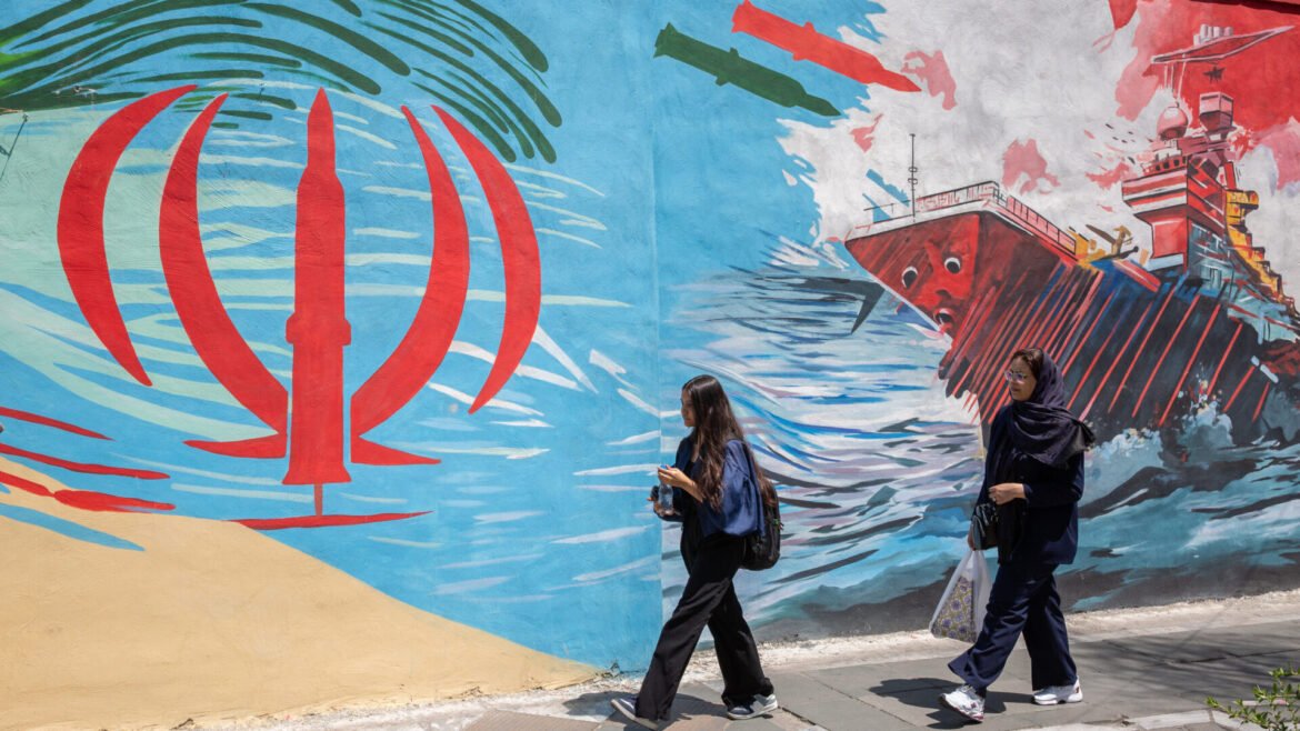 Two women walk past a vibrant mural showing a red ship and blue ocean waves in a coastal scene.