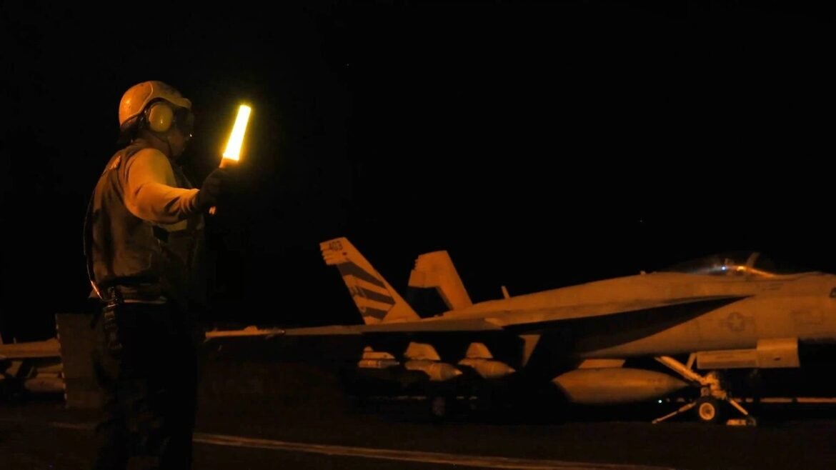 Ground crew member in flight suit and helmet holds a glowing baton on a dark runway beside a fighter jet at night, guiding traffic or signals downrange.