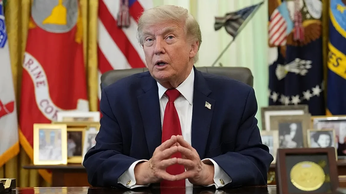 Former U.S. President Donald Trump sits at a desk, hands clasped, in an office with flags and framed photos behind him.