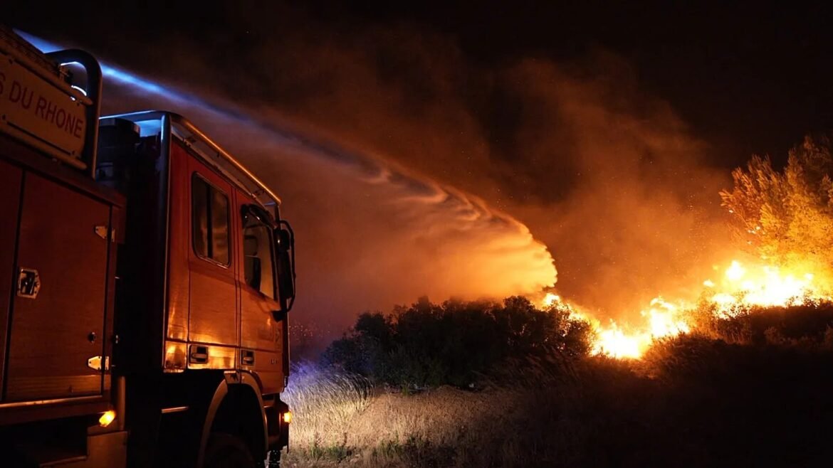 Fire truck at night spraying water toward a grass fire with tall orange flames and smoke behind it.