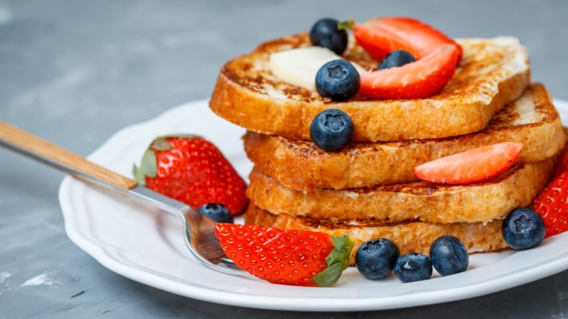 Stack of golden French toast slices on a white plate, topped with strawberries and blueberries and a butter pat nearby.