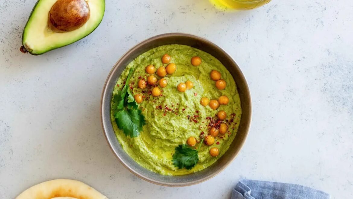 Top-down view of a bowl of vibrant green dip garnished with cilantro leaves and roasted chickpeas, on a light surface.