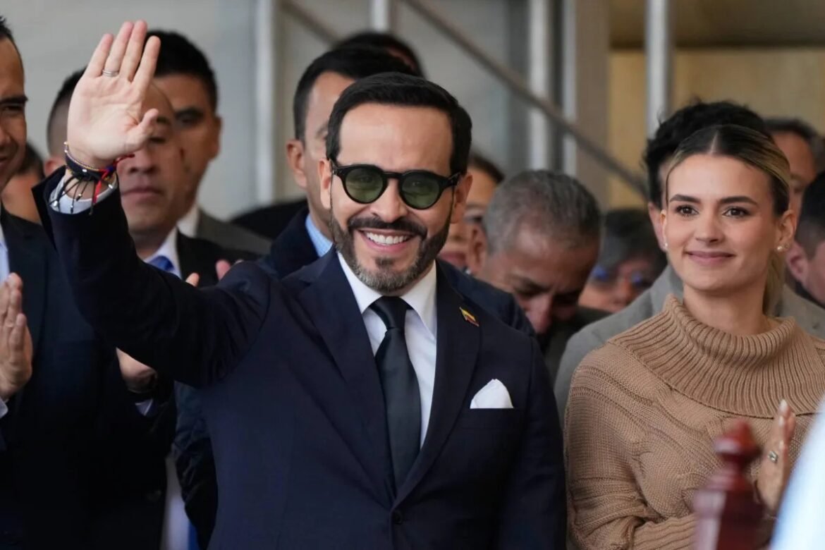 Man in a dark suit and sunglasses waves to a crowd, smiling, with a woman in a tan sweater beside him.
