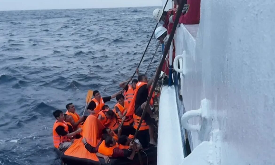 Overcrowded lifeboat with people in bright orange life jackets clinging to a large vessel in rough sea.
