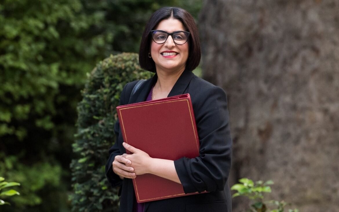 Smiling woman in a black blazer with glasses, holding a red binder outdoors among greenery.