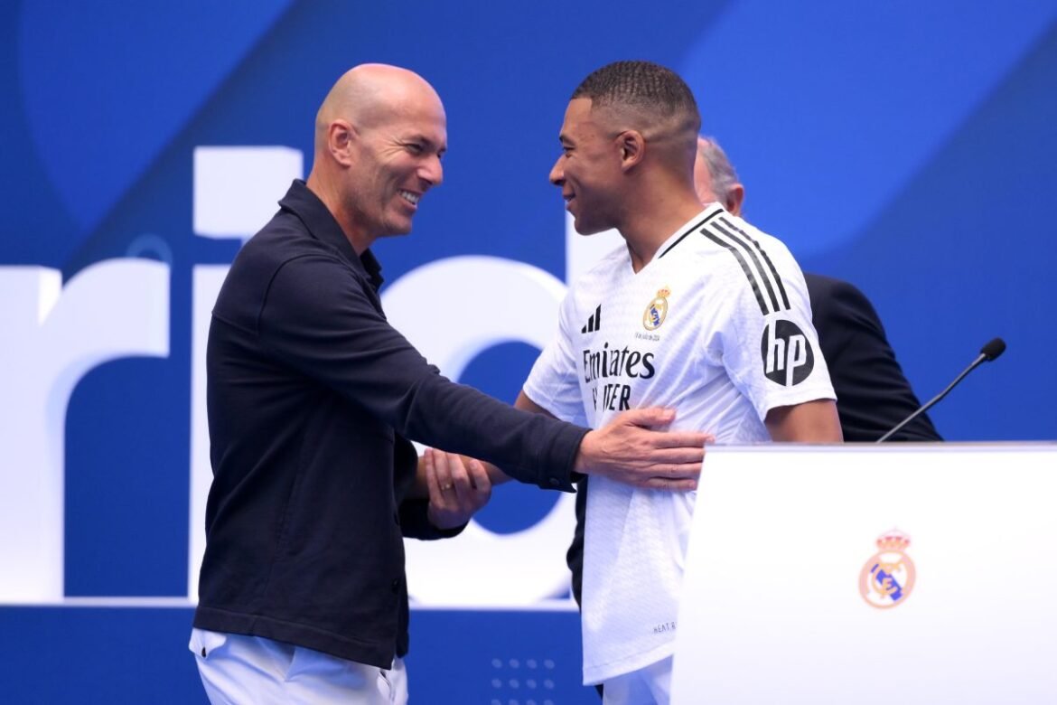 Two men shake hands on a stage, smiling during a Real Madrid event, with one in a white Real Madrid jersey and the other in a dark polo shirt.
