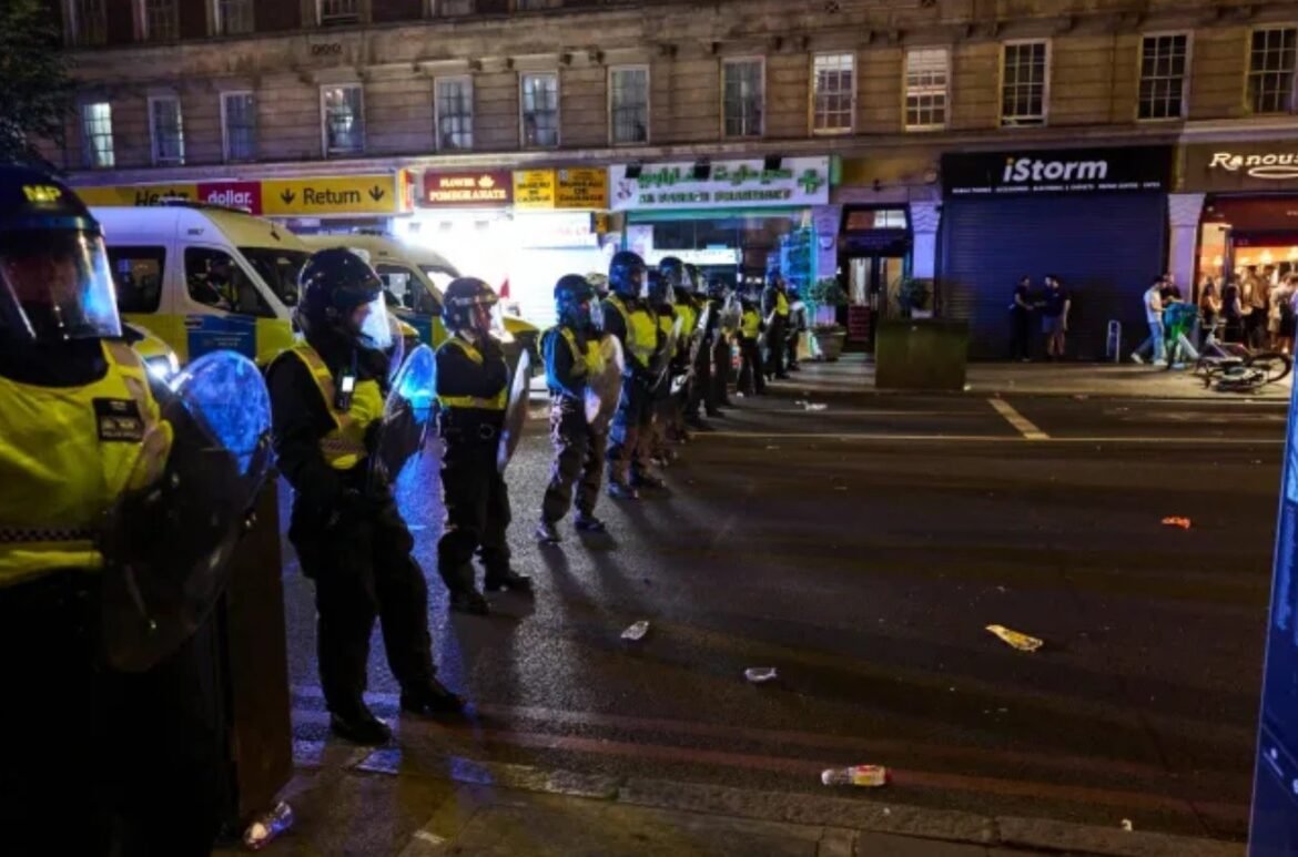 Line of riot police in yellow vests with shields form a barrier along a nighttime city street.
