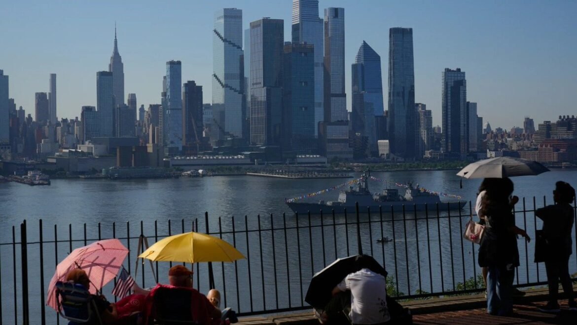 People sit under colorful umbrellas at a railing along the river, with the Manhattan skyline and tall buildings across the water in the background.