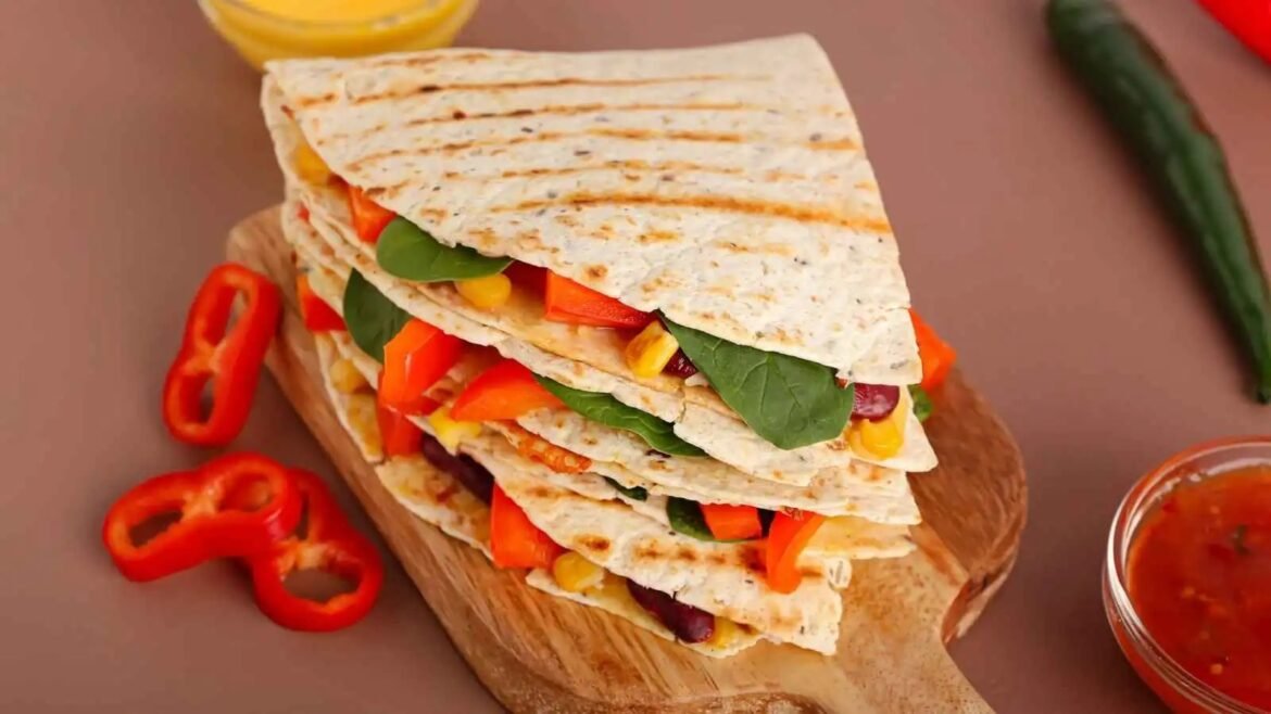 Stacked veggie tortillas with peppers, corn, and greens on a wooden board, ready for dipping in salsa.