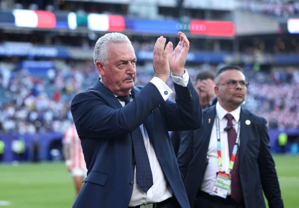 Man in a navy suit applauds on a football field, stadium crowd in the background, another official nearby wearing a lanyard