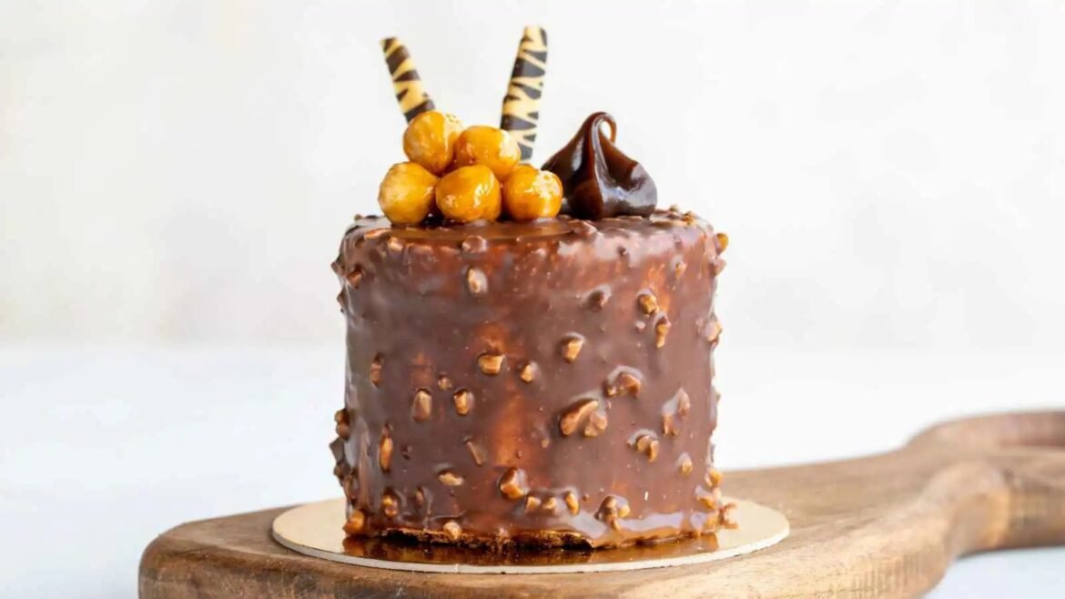 Chocolate cake with glossy chocolate glaze and crushed nuts on a wooden board, topped with caramelized hazelnuts, two striped wafer sticks, and a chocolate swirl attachment.