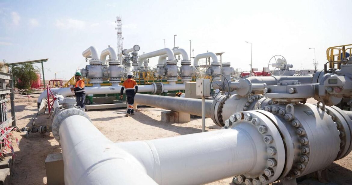 Industrial gas/oil pipeline facility with large white pipes, valves, and support structures; two workers in orange safety vests inspect equipment in a desert site.