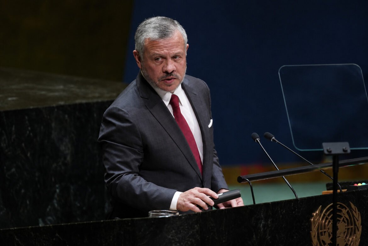 Man in a dark suit and red tie stands at a UN podium with two microphones, gesturing with his hand. Visual of formal speech setting.