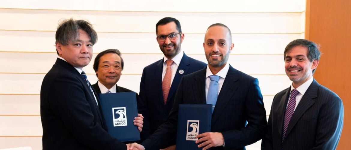 Five men in business suits shake hands during a signing ceremony, each holding blue folders with ADNOC logos.