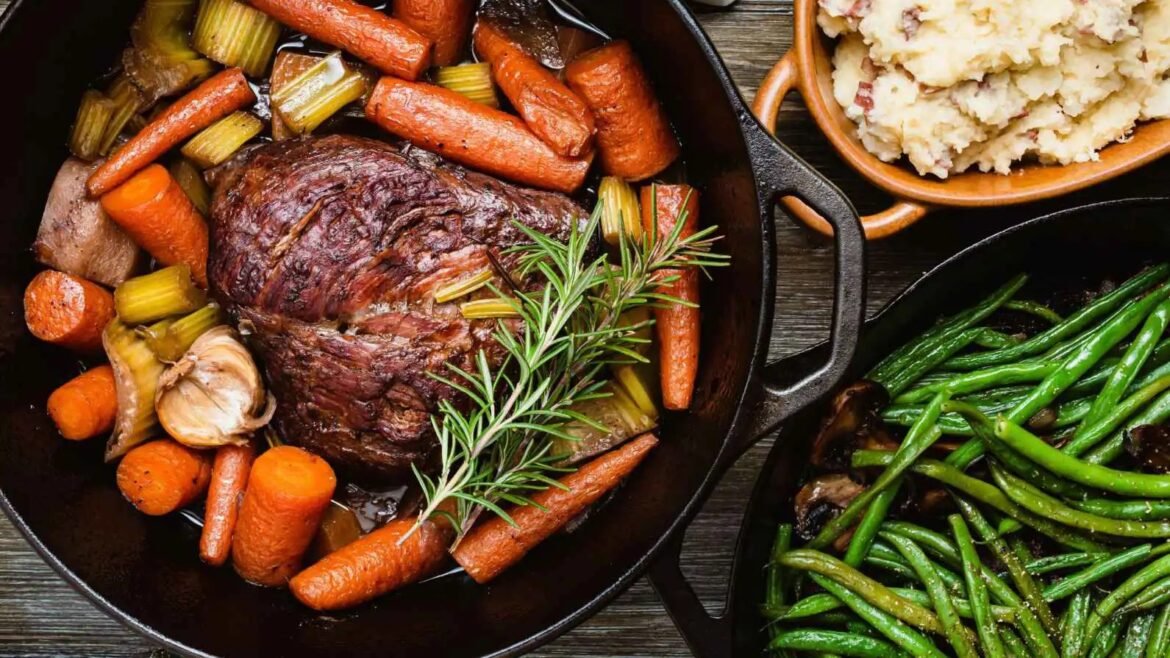 Roast beef with carrots, celery, garlic, and a sprig of rosemary in a cast-iron skillet, with mashed potatoes and green beans nearby.