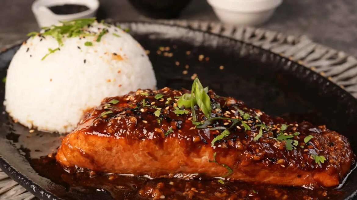 Glazed chicken in a sticky sesame-soy sauce, garnished with herbs, served with steamed white rice on a black plate.