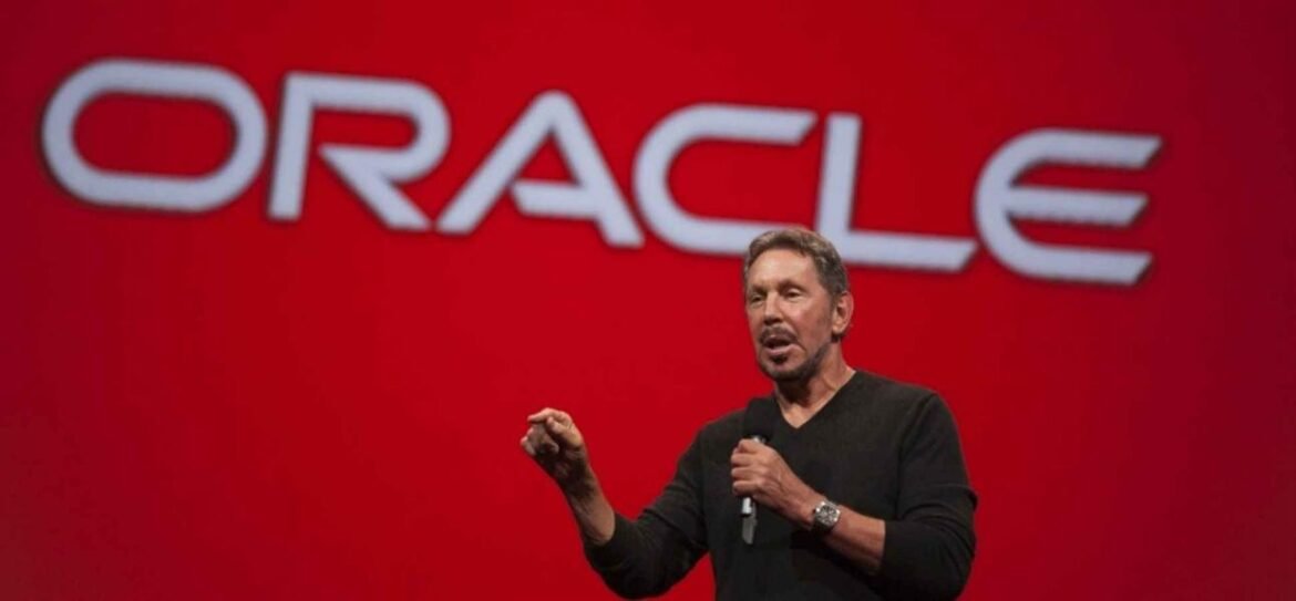 A man on stage speaking into a microphone with a large red backdrop and the Oracle logo behind him.