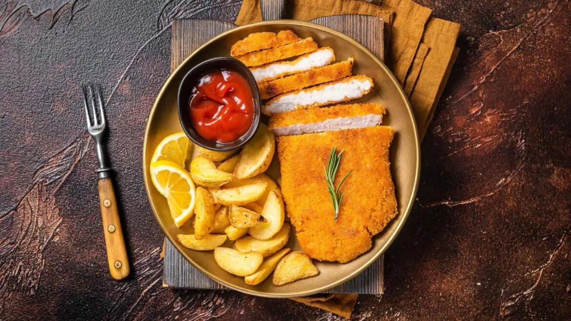 Golden breaded chicken cutlet with potato wedges, lemon slices, and rosemary on a bronze plate.