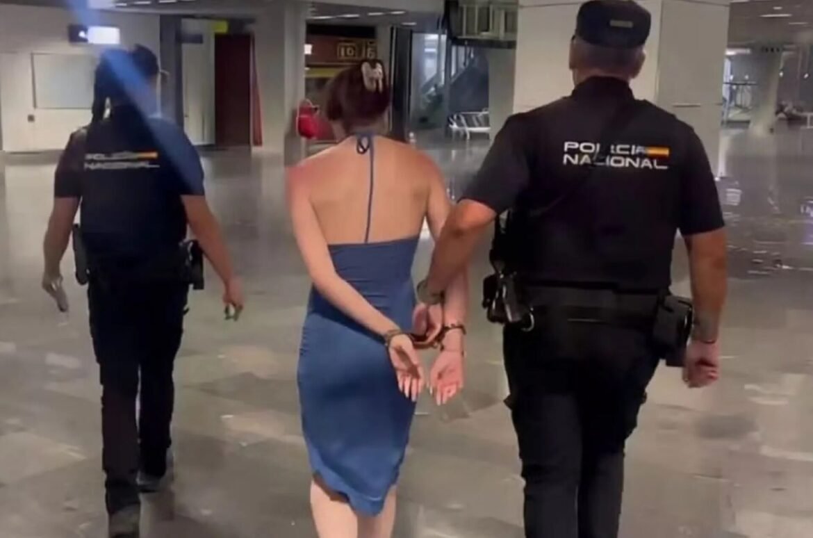 Back view of a woman in a blue dress handcuffed and escorted by two police officers in a hallway.