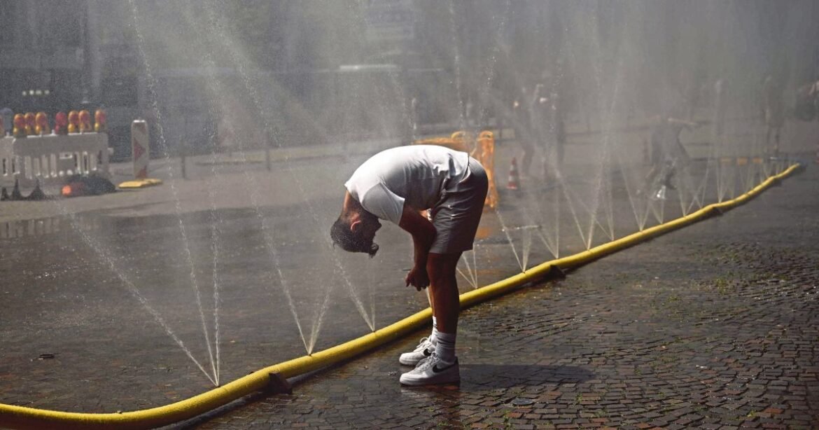 Man bending forward with hands on knees under water sprays emerging from a long yellow hose on a cobblestone street.