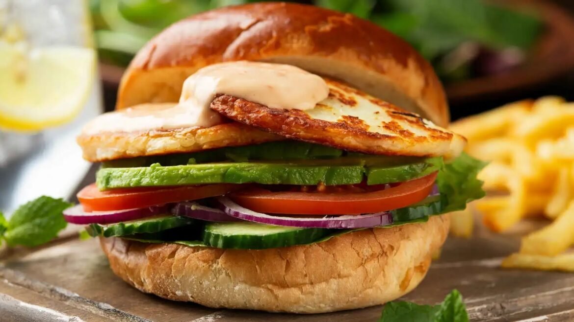 Burger with grilled patty, avocado, tomato, red onion, cucumber and lettuce on a toasted bun with creamy sauce on top.