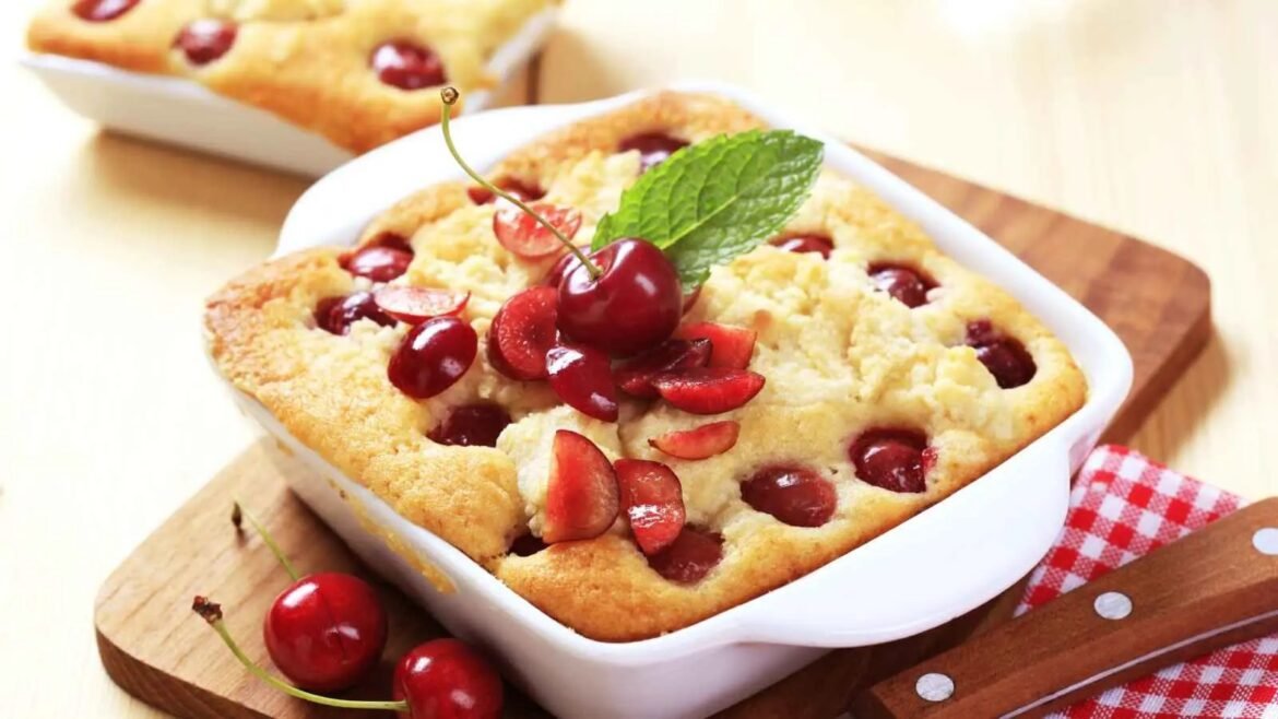 Cherry clafoutis or crumble in a white baking dish, topped with fresh cherries and a mint leaf on a wooden board.