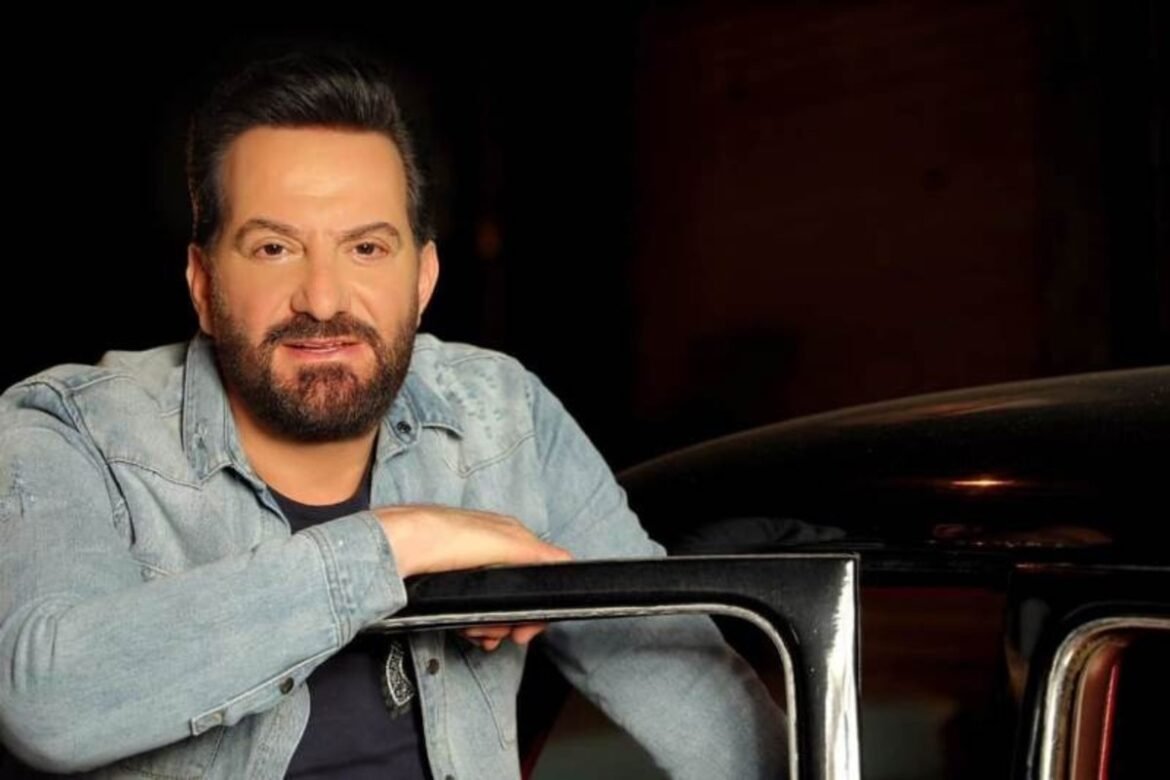 Man with dark hair and beard in a light denim jacket leans on a car door at night, smiling slightly.