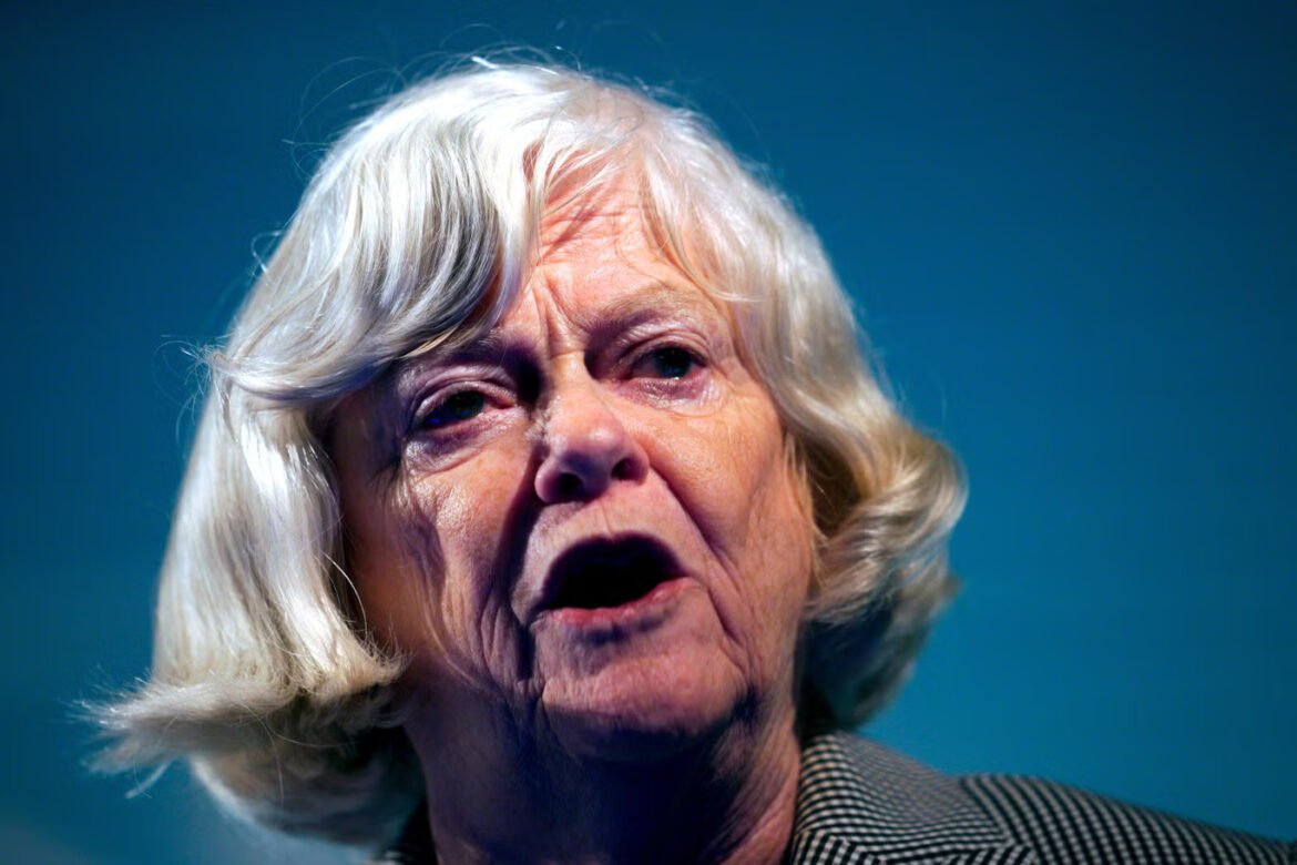 Close-up of an elderly woman with white hair speaking, mouth open, wearing a checkered blazer against a blue background.