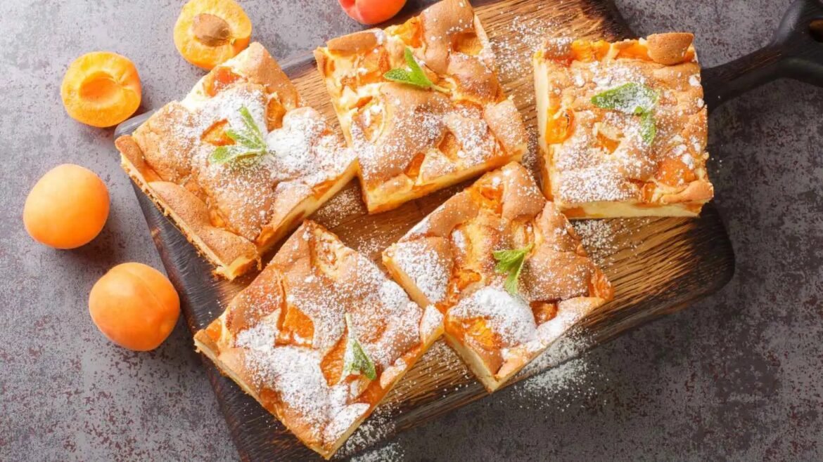 Rectangular apricot bars dusted with powdered sugar on a dark wooden board, garnished with mint leaves and fresh apricots nearby.