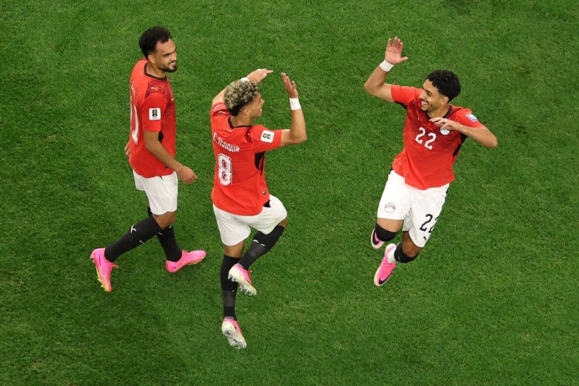 Three soccer players in red jerseys celebrate on a green field, jumping and high-fiving each other.