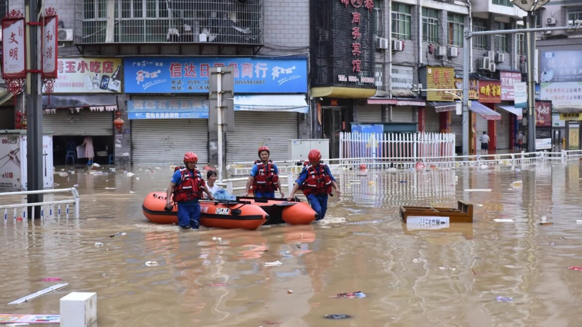 Rescue workers in red life jackets and helmets push an orange inflatable boat through a flooded urban street with shuttered shops behind them.