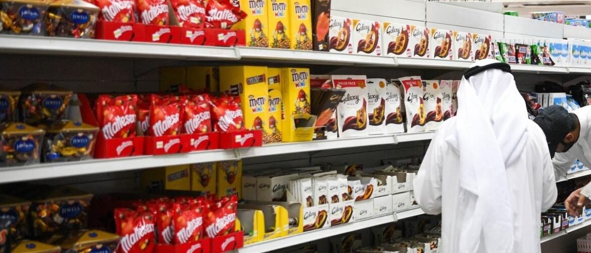 A man in traditional white attire browses chocolate boxes on store shelves in a supermarket aisle.