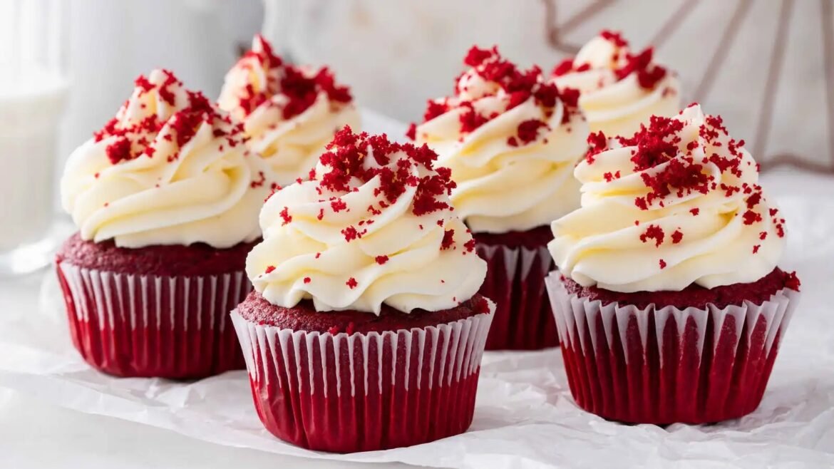 Red velvet cupcakes topped with swirls of white cream cheese frosting and red crumb sprinkles on a parchment-lined surface.