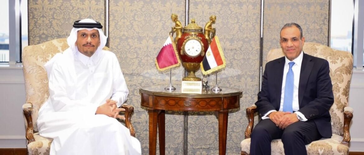 Two men sit in ornate armchairs with a decorative table between them, a clock and small flags in front, suggesting a formal meeting.