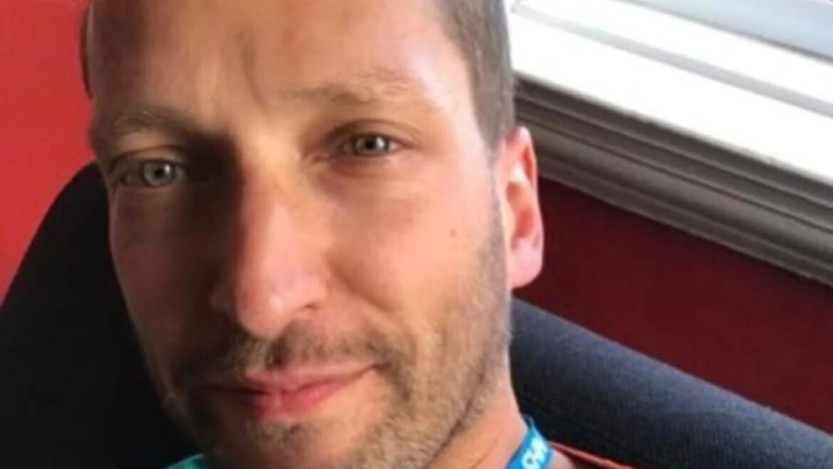 Close-up of a smiling man with light stubble, wearing a blue lanyard, indoors near a red wall and window frame.