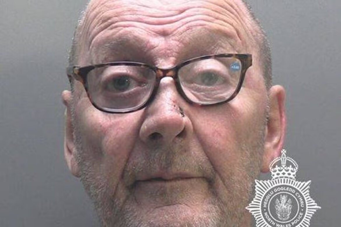 Mugshot of an older man with glasses against a gray backdrop, police badge watermark in the corner.