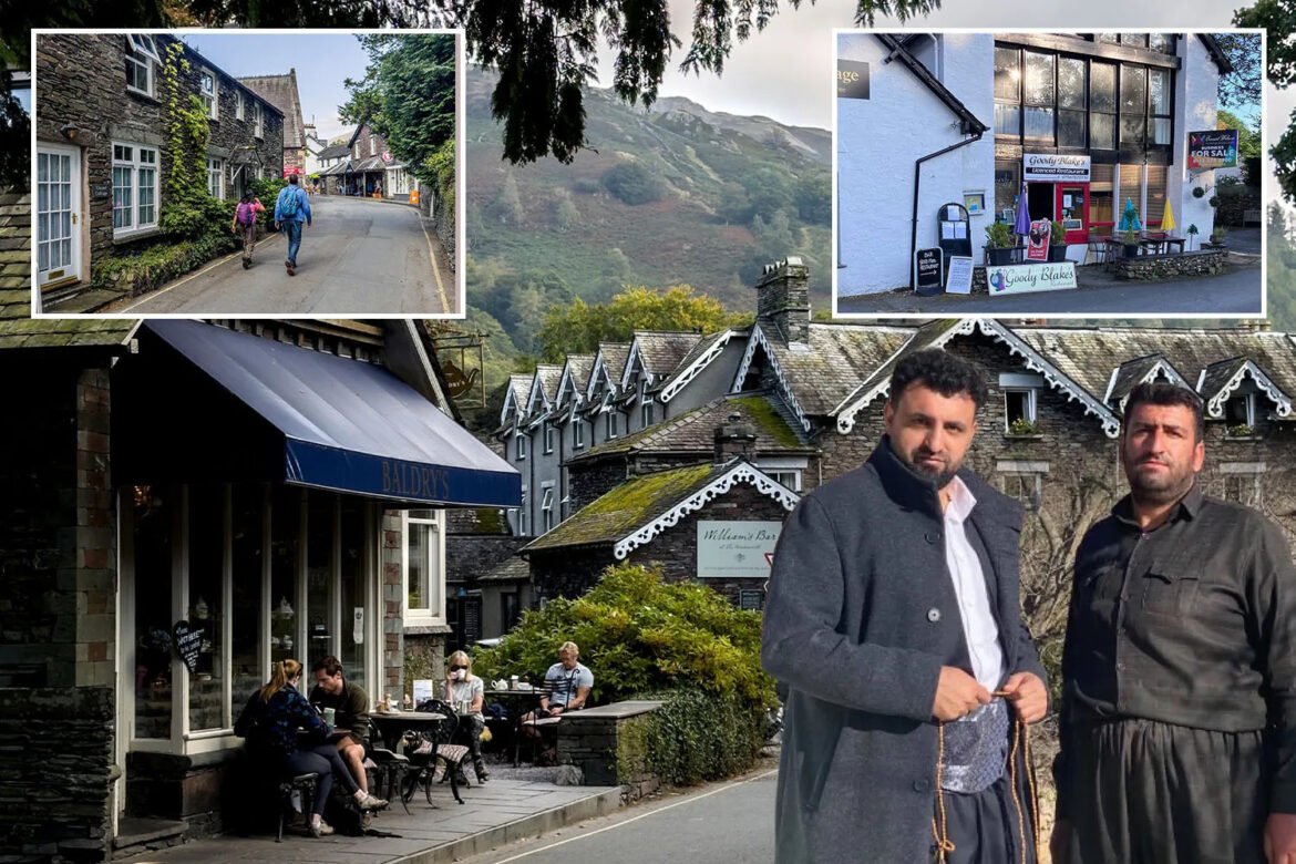 Two men in coats stand in the foreground of a stone village street with an outdoor café to the left and hills in the background.