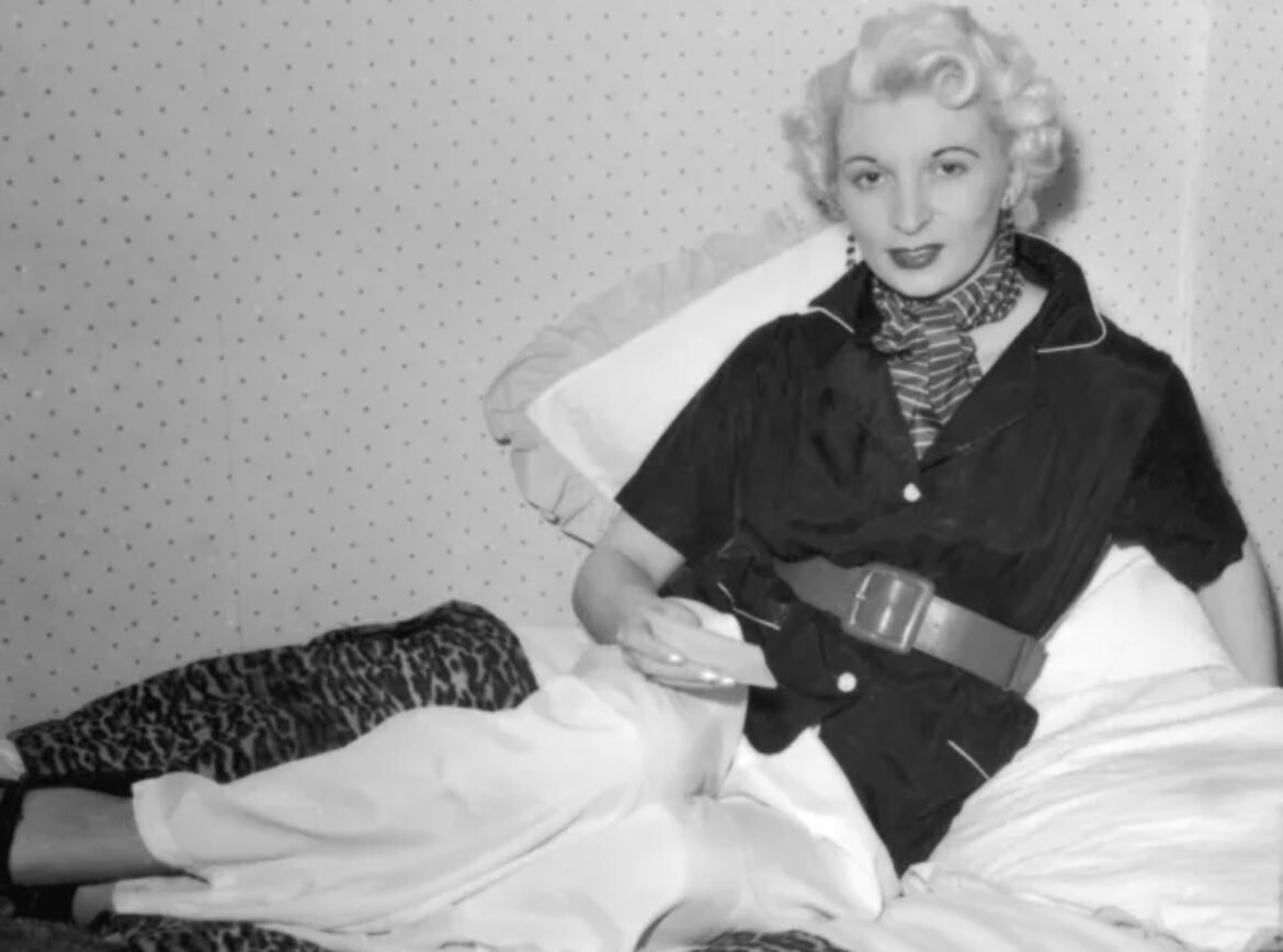 Black-and-white photo of a vintage-style woman posing on a bed, wearing a belted robe and scarf with curled hair.