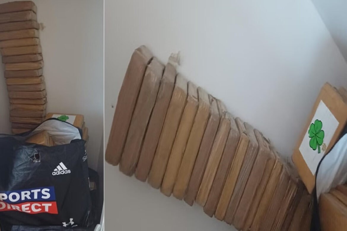 Corner of a room with a tall stack of flat wooden boards leaning upright against a white wall, next to a black Sports Direct bag and a framed shamrock drawing.