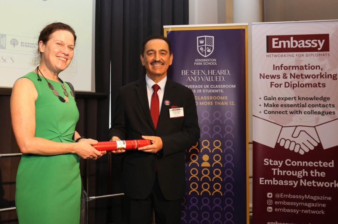 Two adults exchange a red diploma or scroll at a formal event, with banners in the background promoting education and diplomacy networks.