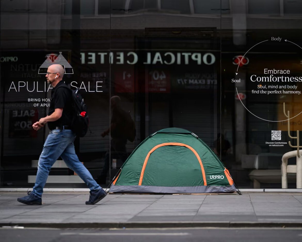 Man in a black shirt and jeans walks past a small green tent with orange trim on a city sidewalk.