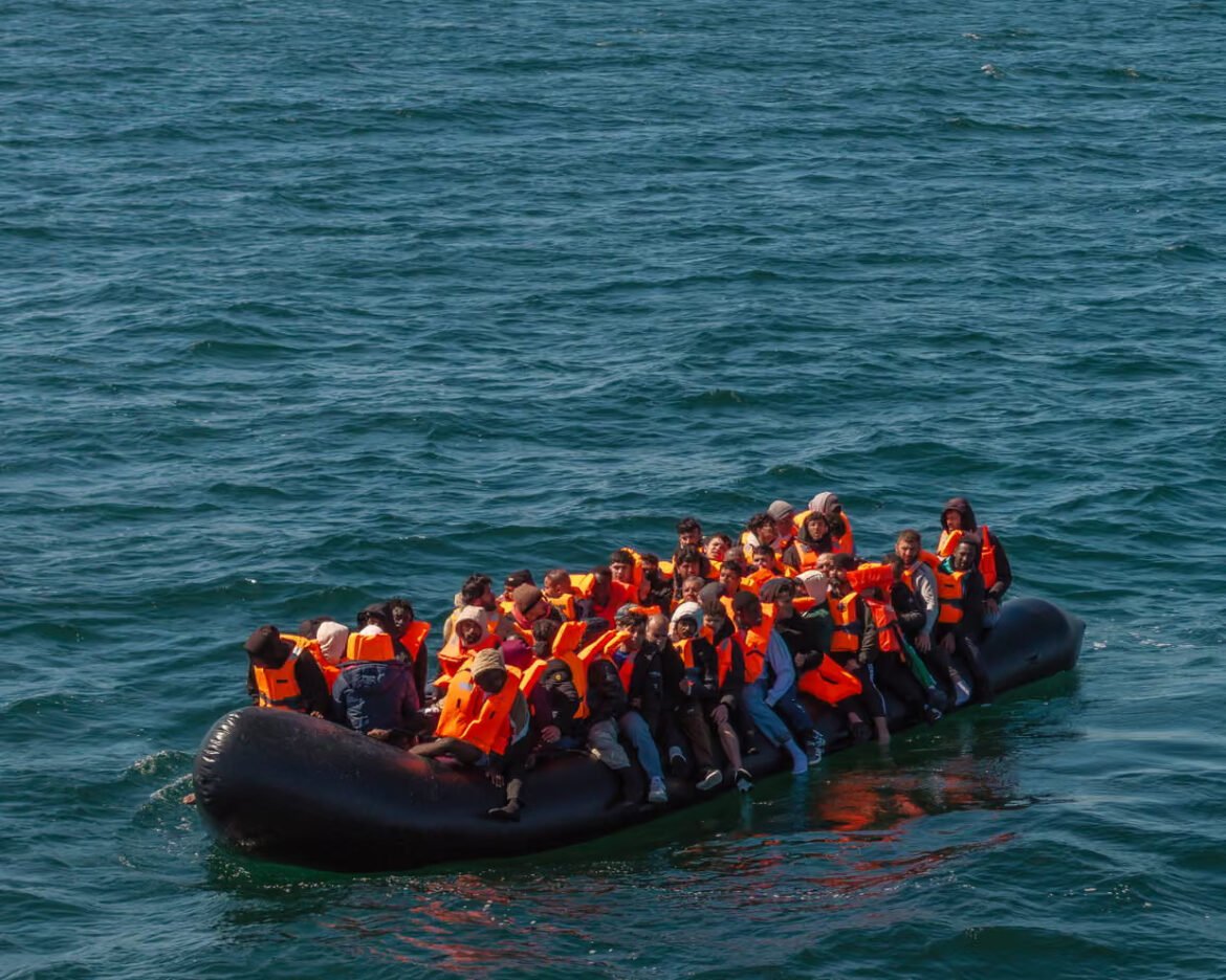 Crowded inflatable boat filled with people wearing bright orange life jackets on blue open water.