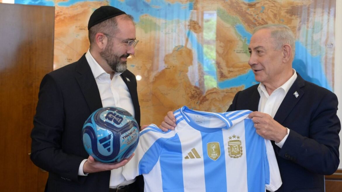 Two men in suits stand by a world map; one wearing a kippah shows an Argentina jersey to the other while holding a blue soccer ball.