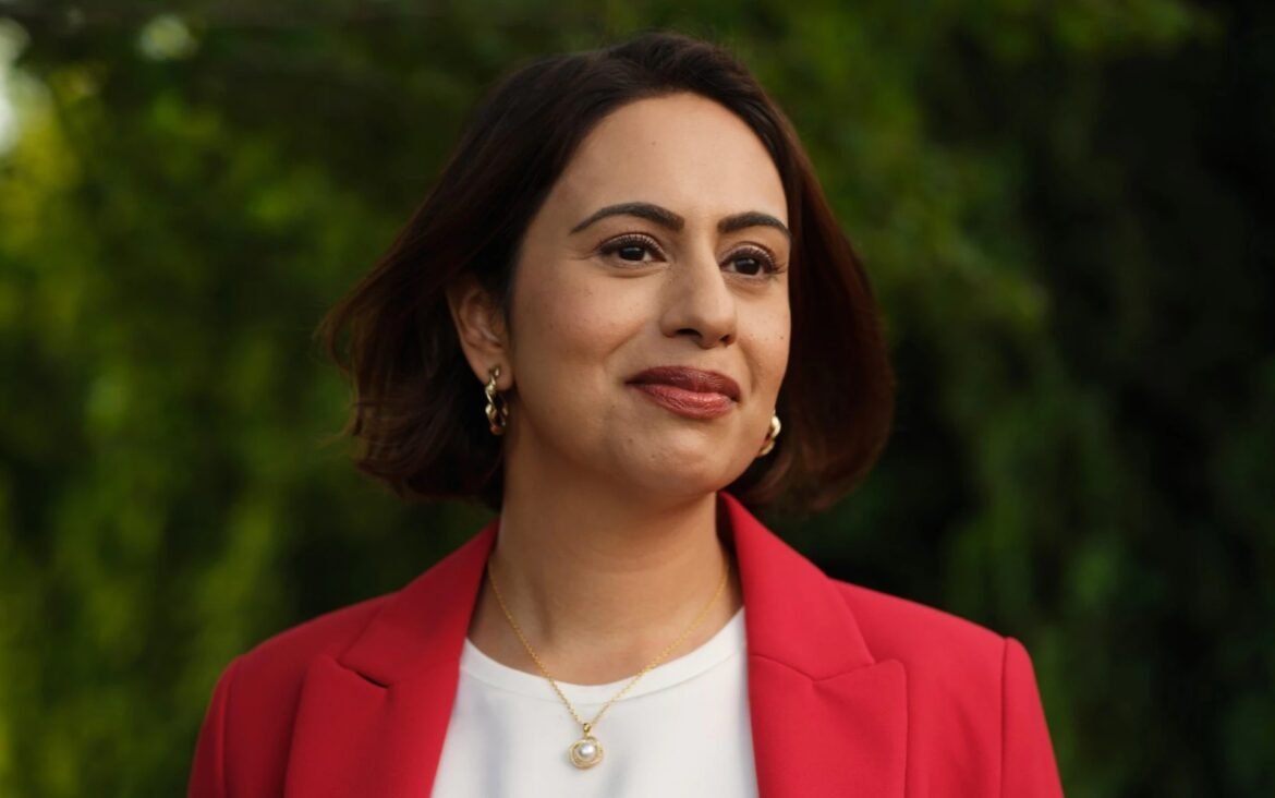 Portrait of a woman with short dark hair wearing a red blazer and gold jewelry, outdoors with a green blurred background, smiling slightly to the left.