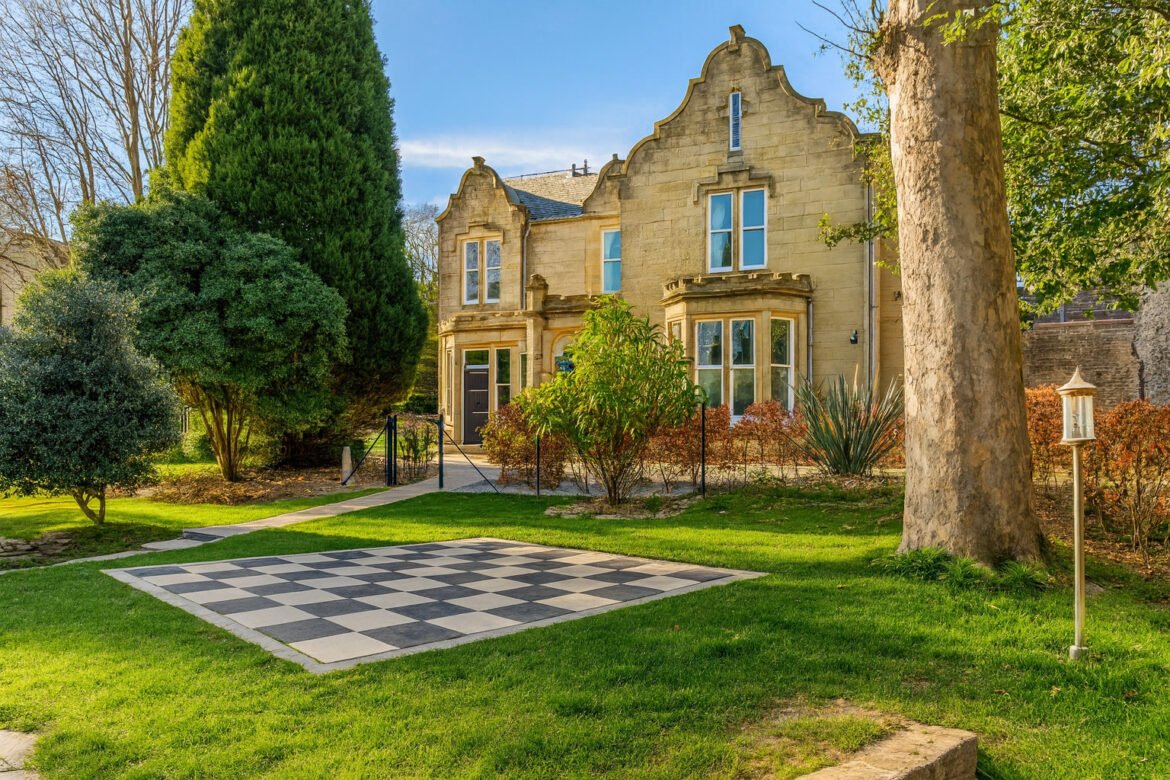 Stone two-story house with ornate gables visible behind a green lawn and shrubbery, next to a large outdoor checkerboard.