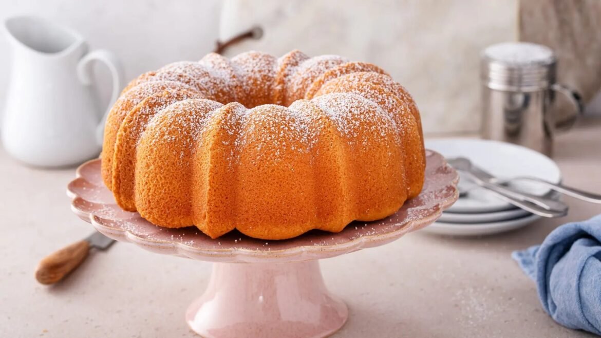 Bundt cake dusted with powdered sugar sits on a pink cake stand, with a white pitcher and plates in the background.