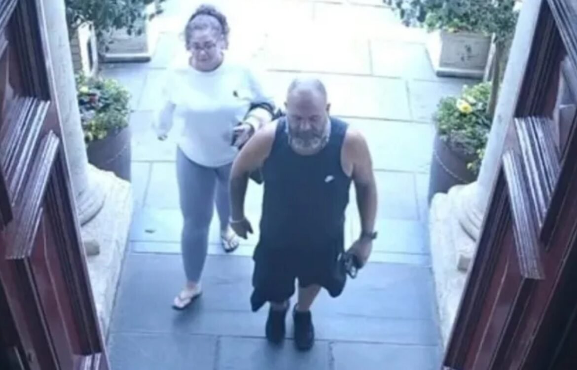 Two adults walk up a front porch staircase toward the camera; man in a black sleeveless shirt leads, holding sunglasses, with a woman in a light top following behind.