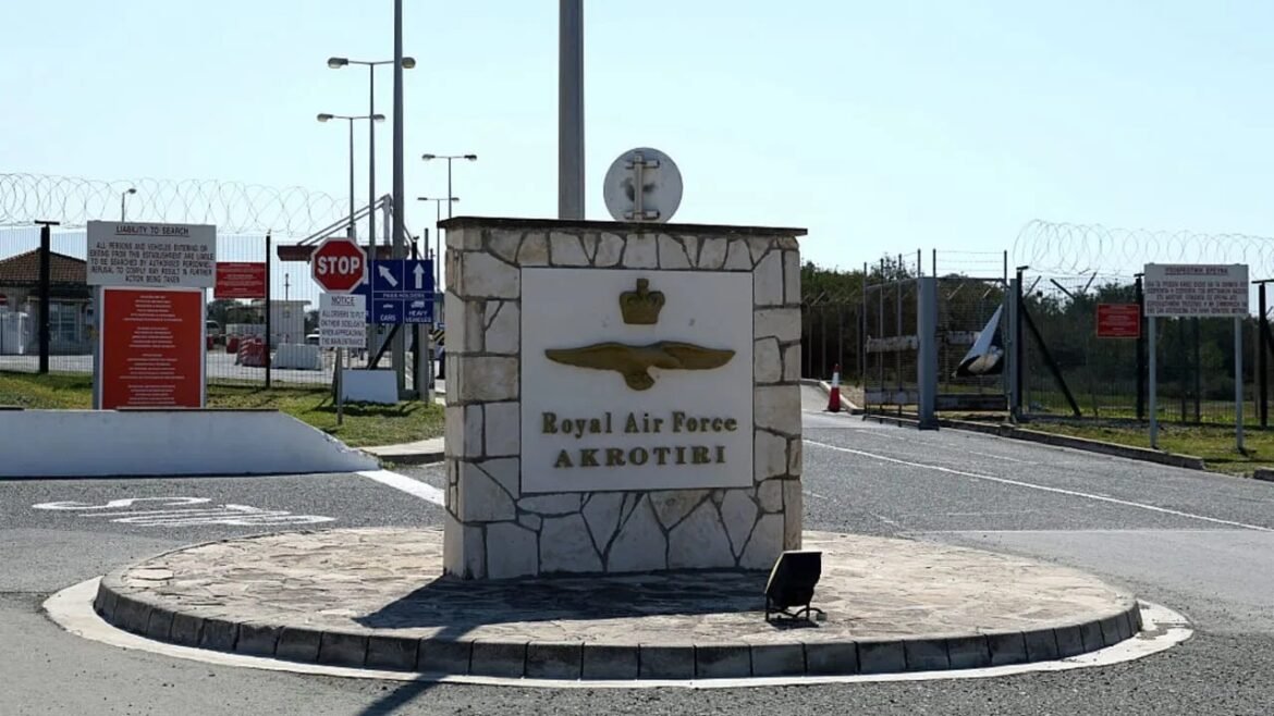 Stone entrance sign reading 'Royal Air Force Akrotiri' with a gold wing emblem at a roundabout. Barbed fence and security signs in the background.