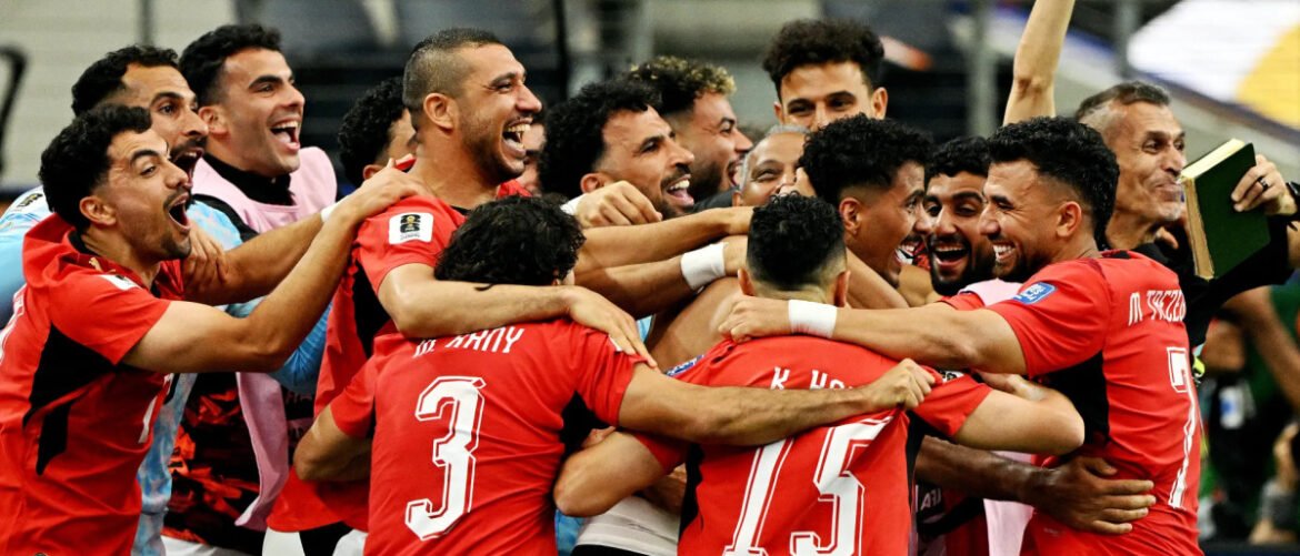 A group of players in red jerseys huddle together, arms around each other, celebrating with smiles after a win.